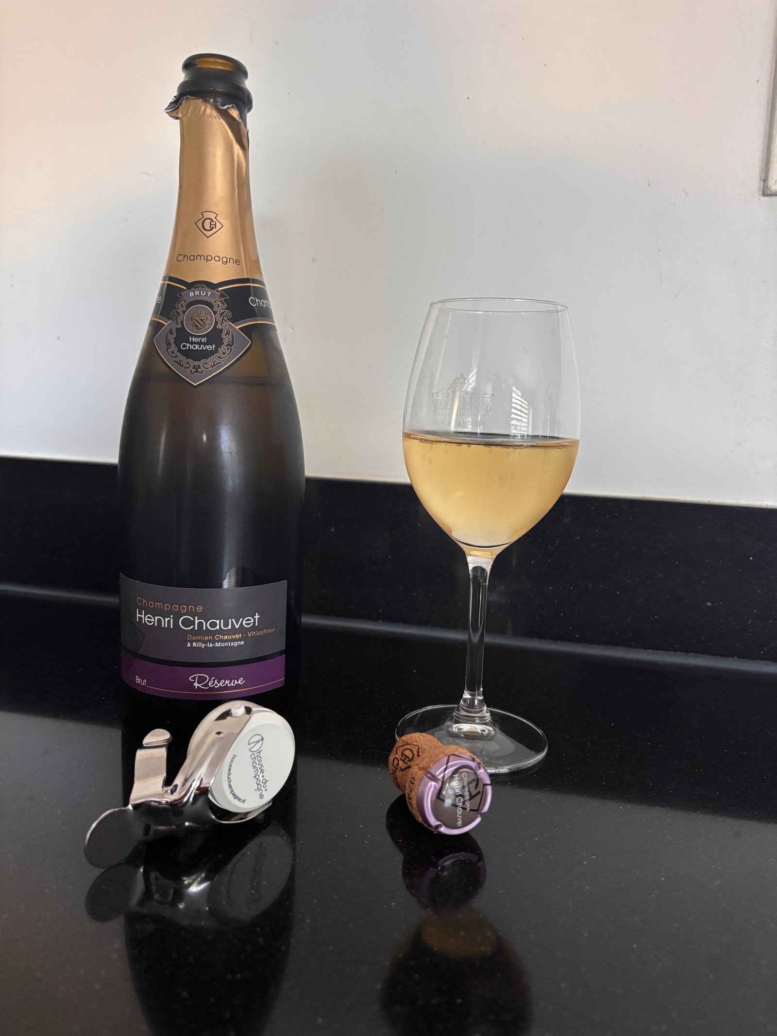 A bottle of Henri Chauvet Brut Réserve Champagne beside a poured glass, cork and stopper on a kitchen counter